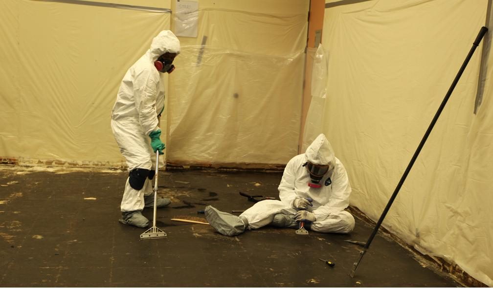 Two workers in protective suits and respirators removing material inside a sealed containment area