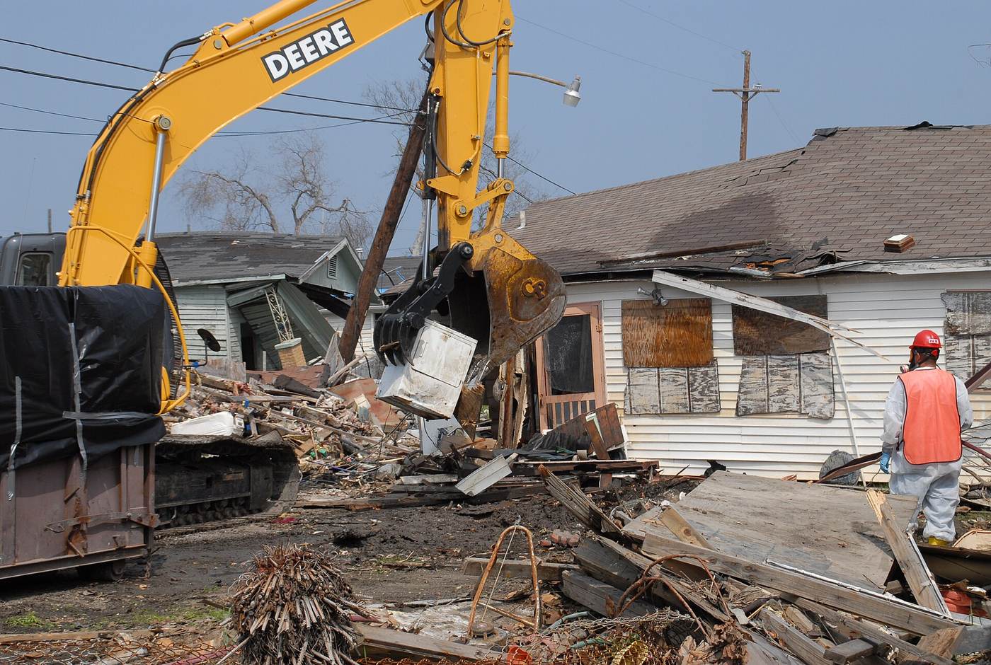 An excavator pulling apart a wood-frame house while a worker in a safety vest watches